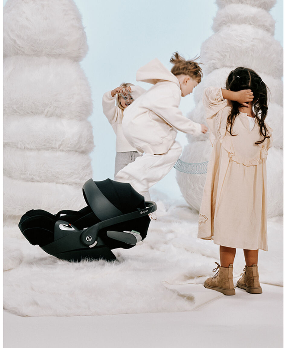 Trois enfants jouant sur un tapis blanc qui ressemble à de la neige. À leur gauche se trouve une coque auto pour bébé CYBEX.
