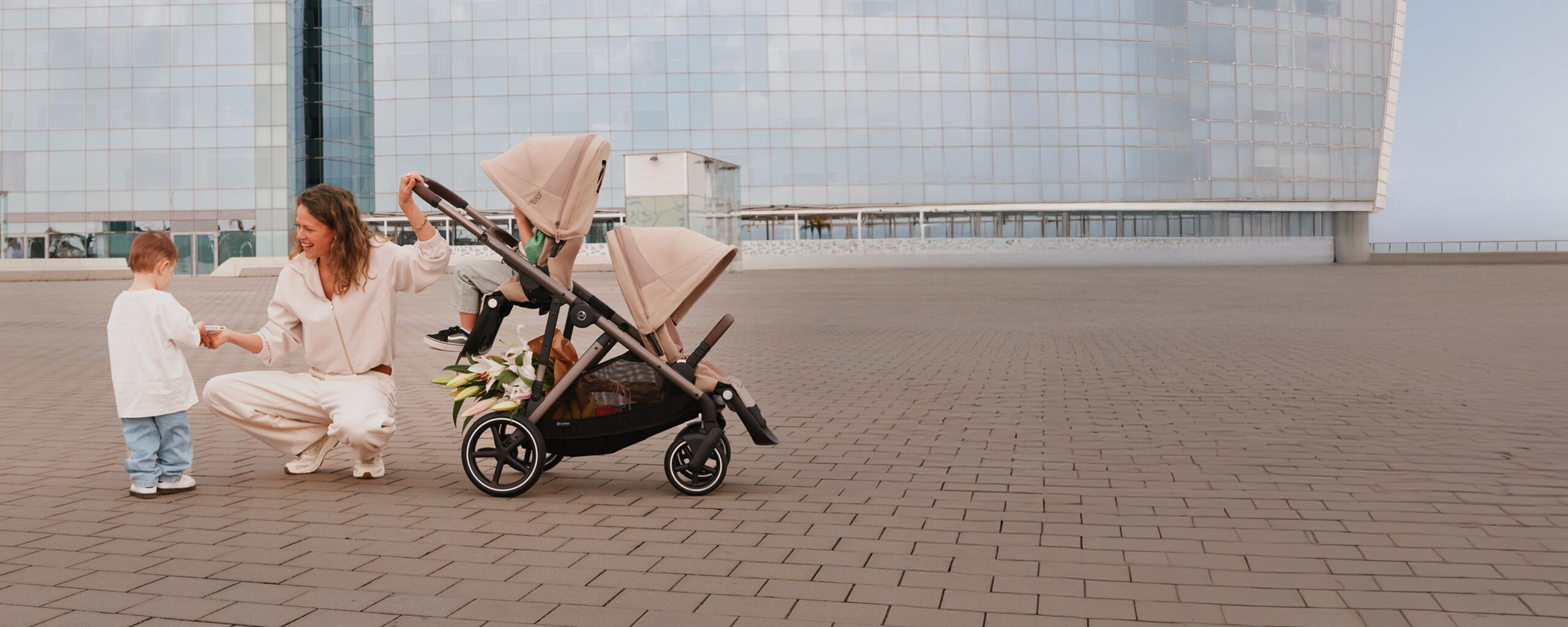 A woman laughing as she kneels down and hands something to her son, one hand on the Gazelle S stroller's handlebar with one child sitting in one of the seat units attached.