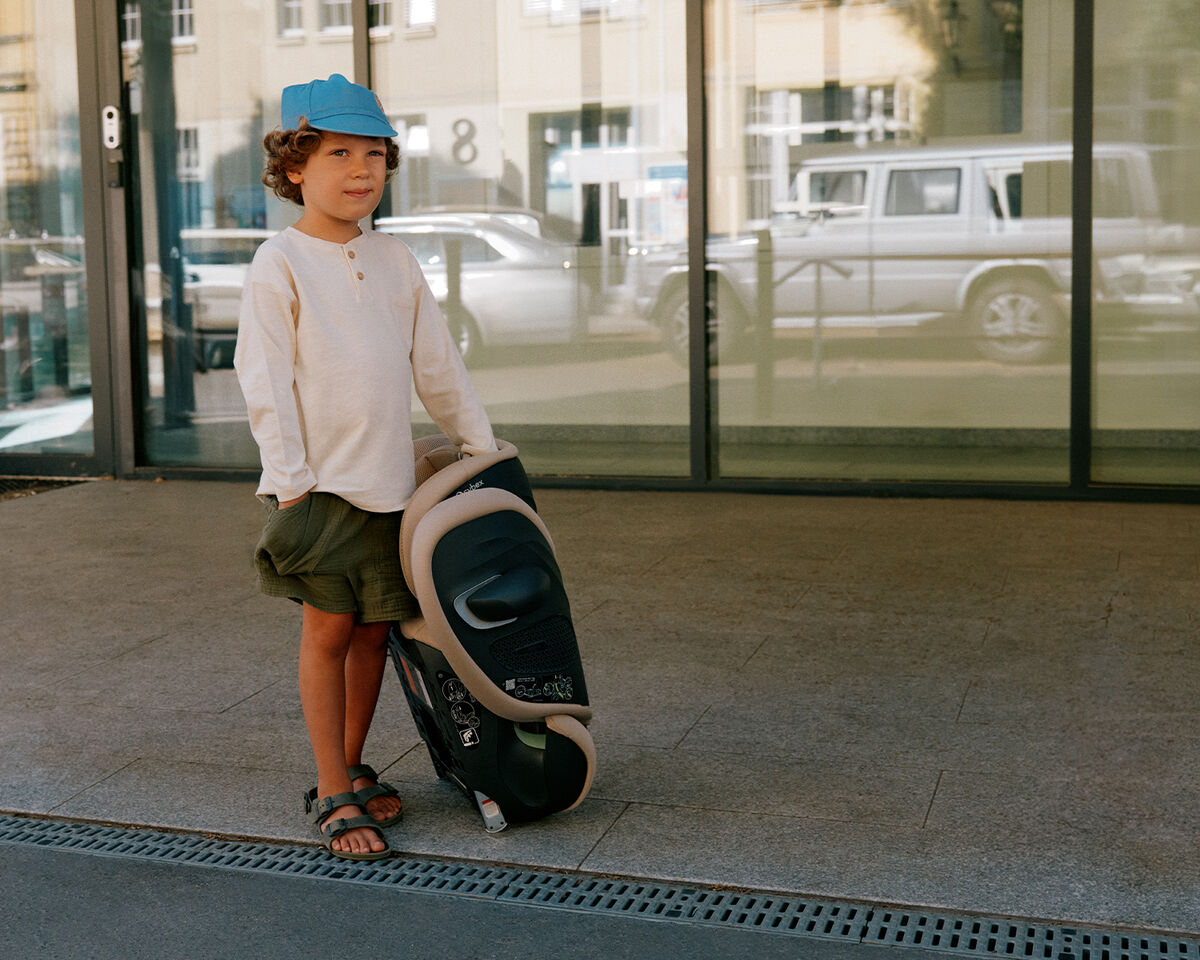 A boy with curly hair stands in front of a modern building holding a stroller.