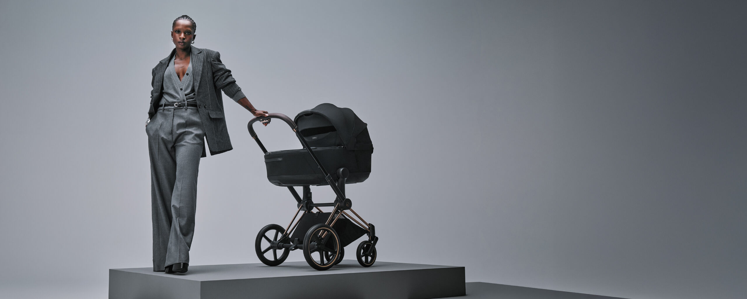 A woman wearing all gray with one hand in her pants pocket and the other holding the handlebar of her Priam stroller.