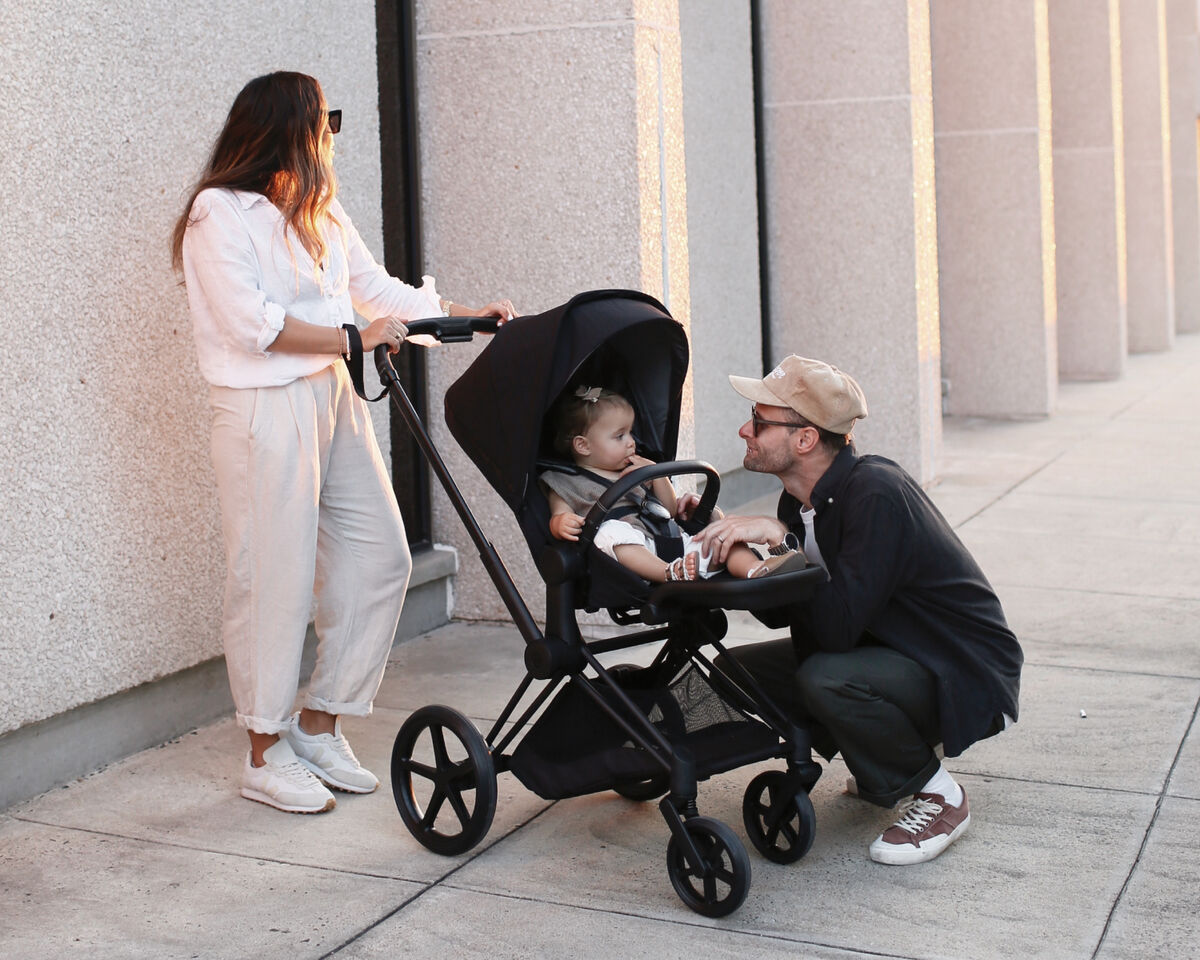Woman in sunglasses pushing a pram with a small child, while a man crouches and talks to the child.