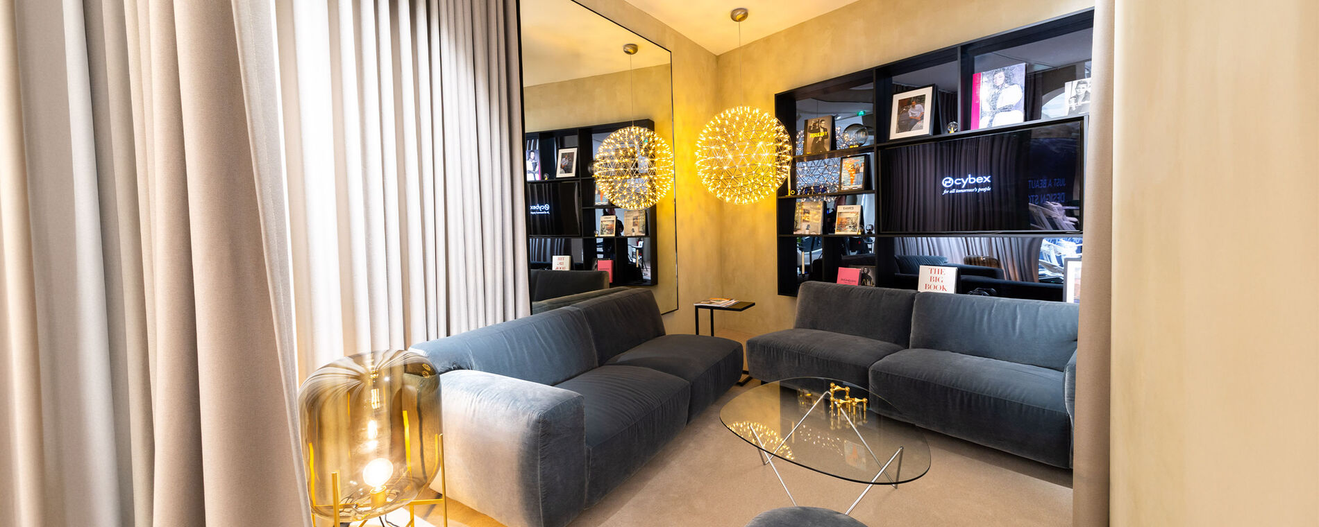 CYBEX store interior with two anthracite sofas placed at a 90-degree angle, an elegant black bookshelf and a sculptural molecule-style floor lamp. A glass coffee table sits between the sofas, with reflections enhancing the luxurious atmosphere.