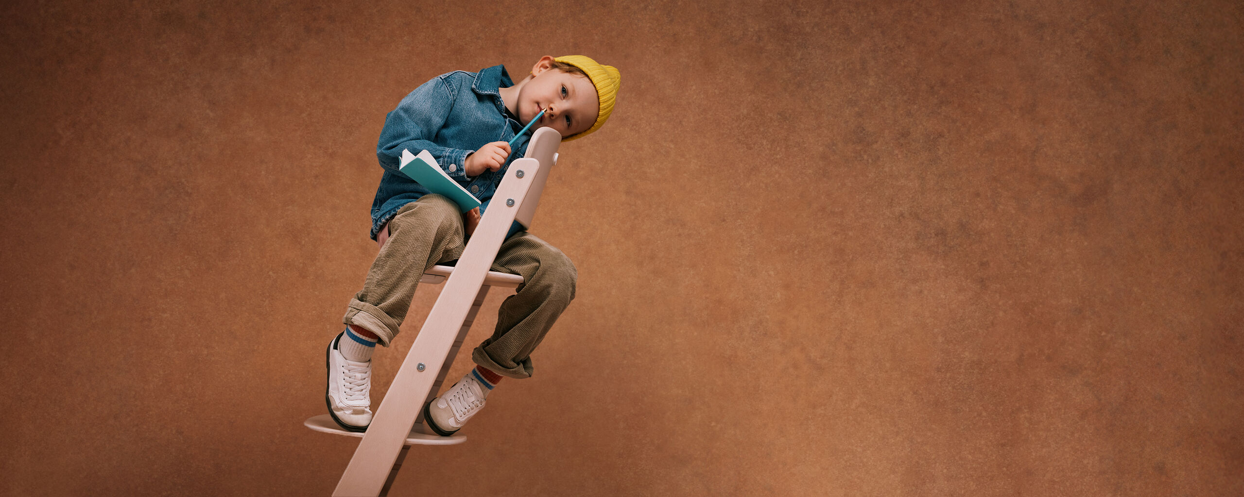 Young child sitting in the CYBEX Iris high chair, head leaning against the backrest with a pen against his lips and a notebook in his hand.
