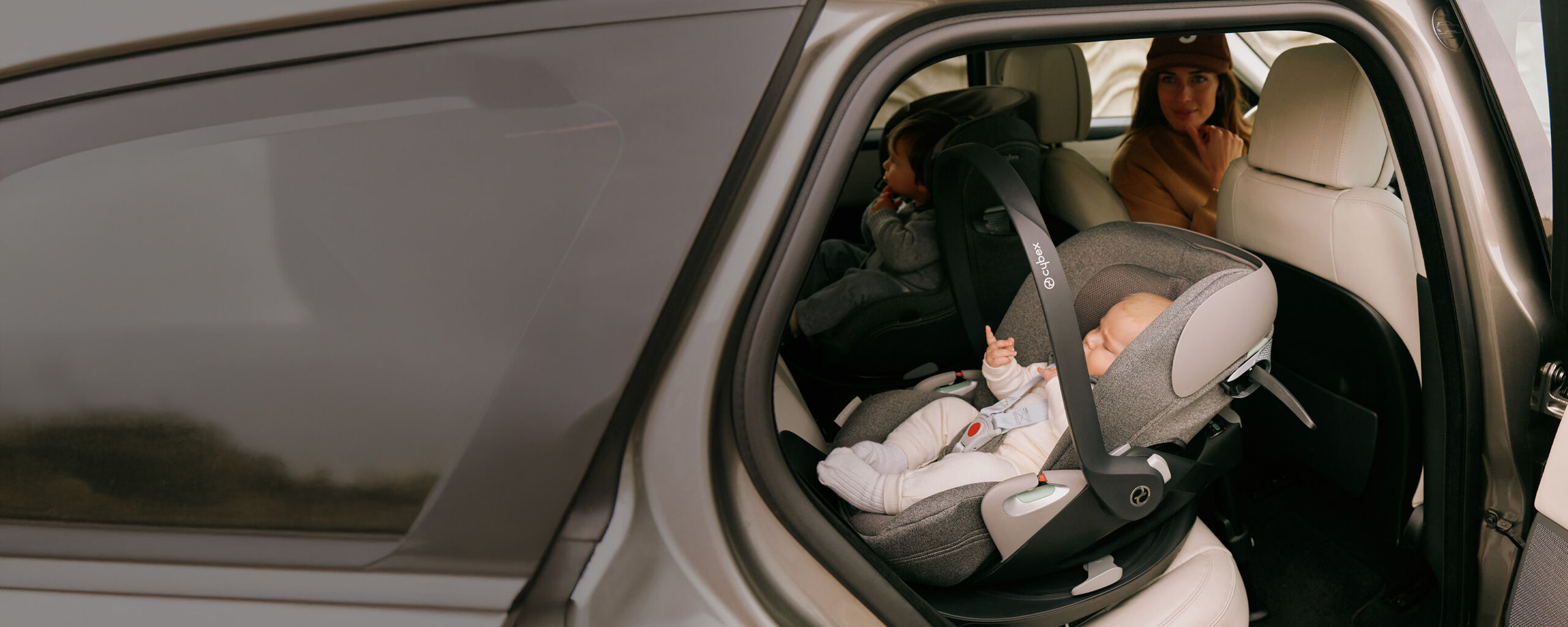 Durch die ge&ouml;ffnete Autot&uuml;r: Ein Baby in einer grauen CYBEX Babyschale reist r&uuml;ckw&auml;rtsgerichtet, w&auml;hrend eine Frau am Fahrersitz nach hinten schaut &ndash; Sicherheit und Kontrolle auf jeder Fahrt.