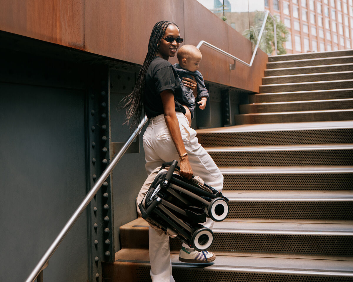 Woman wearing sunglasses carrying a toddler and a folded pushchair up a staircase.