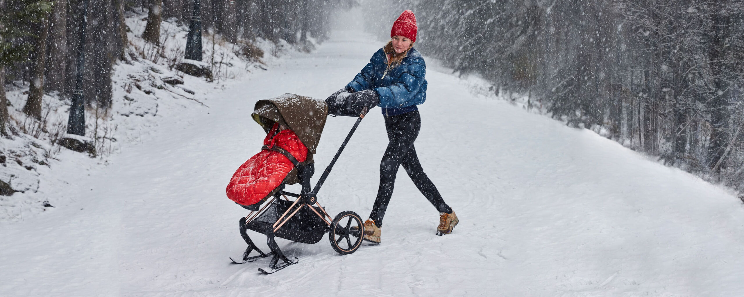 A woman pushes a pram in the snow while it's snowing. She is wearing a red hat and a blue jacket.