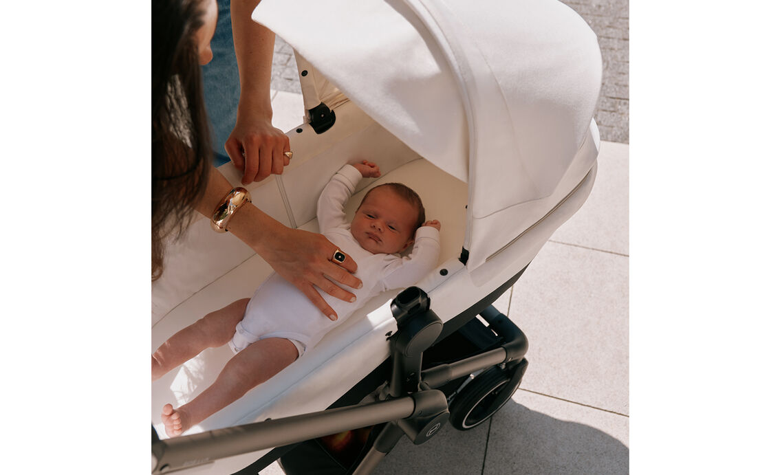 A newborn lies in a CYBEX stroller with Seashell Beige fabric. The mother leans over from above, her hand gently stroking the baby.