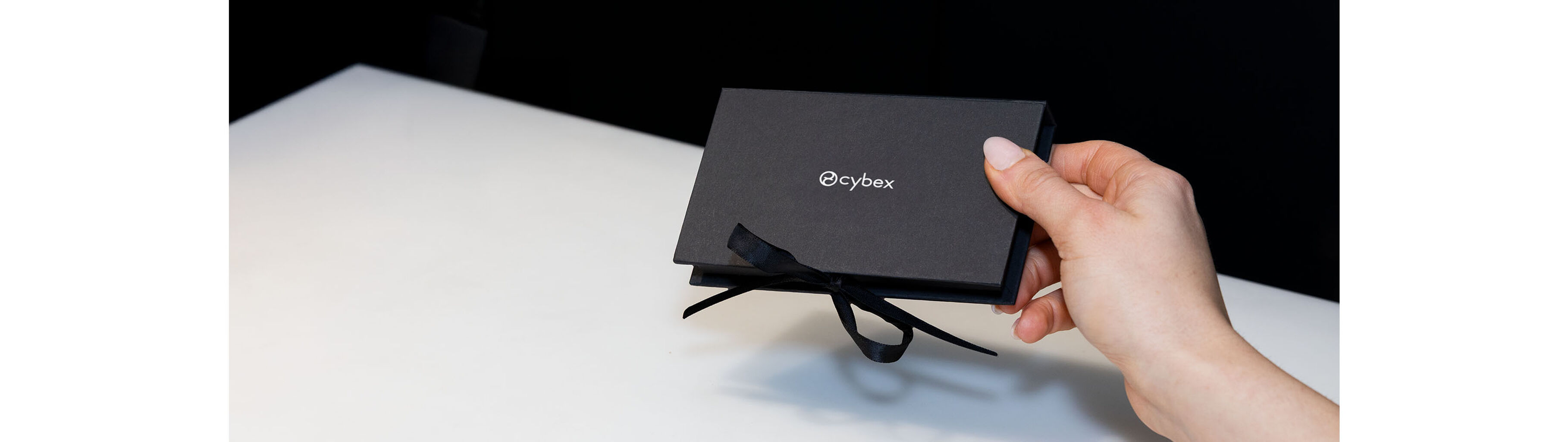 Detail shot: hand with pale pink manicure handing over a black gift box with silver CYBEX logo and black ribbon.