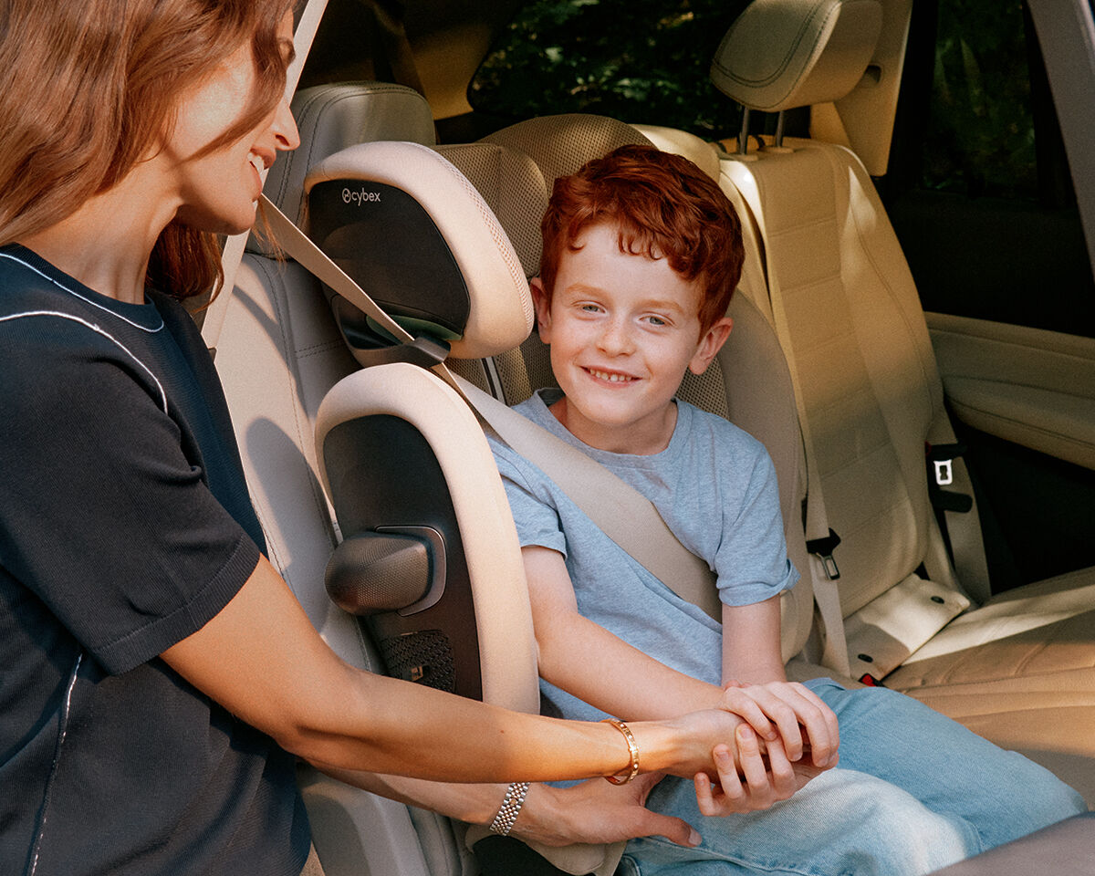 Solution G2 car seat, a smiling boy securely buckled in a car, while a woman gently holds his hand.