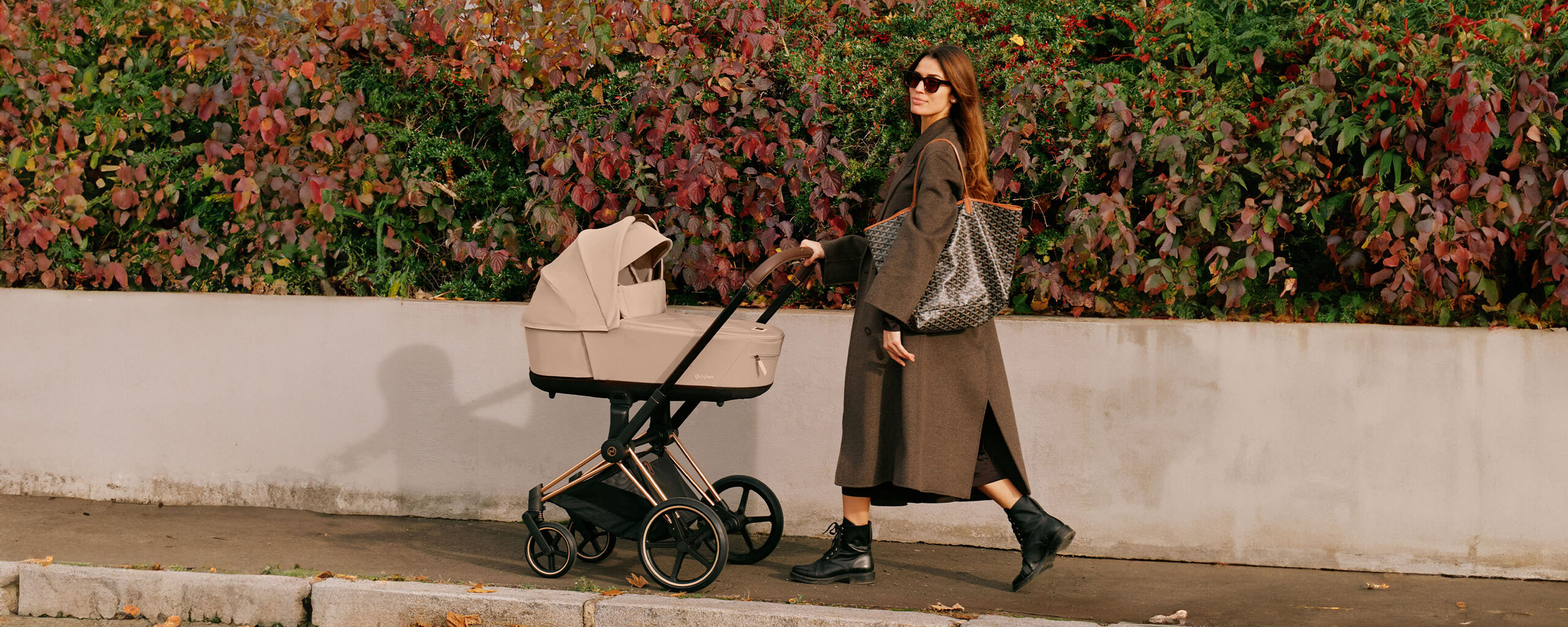 A woman pushes a beige pram along a pavement in front of an autumnal hedge. She is wearing a long coat, boots, and carrying a large bag.