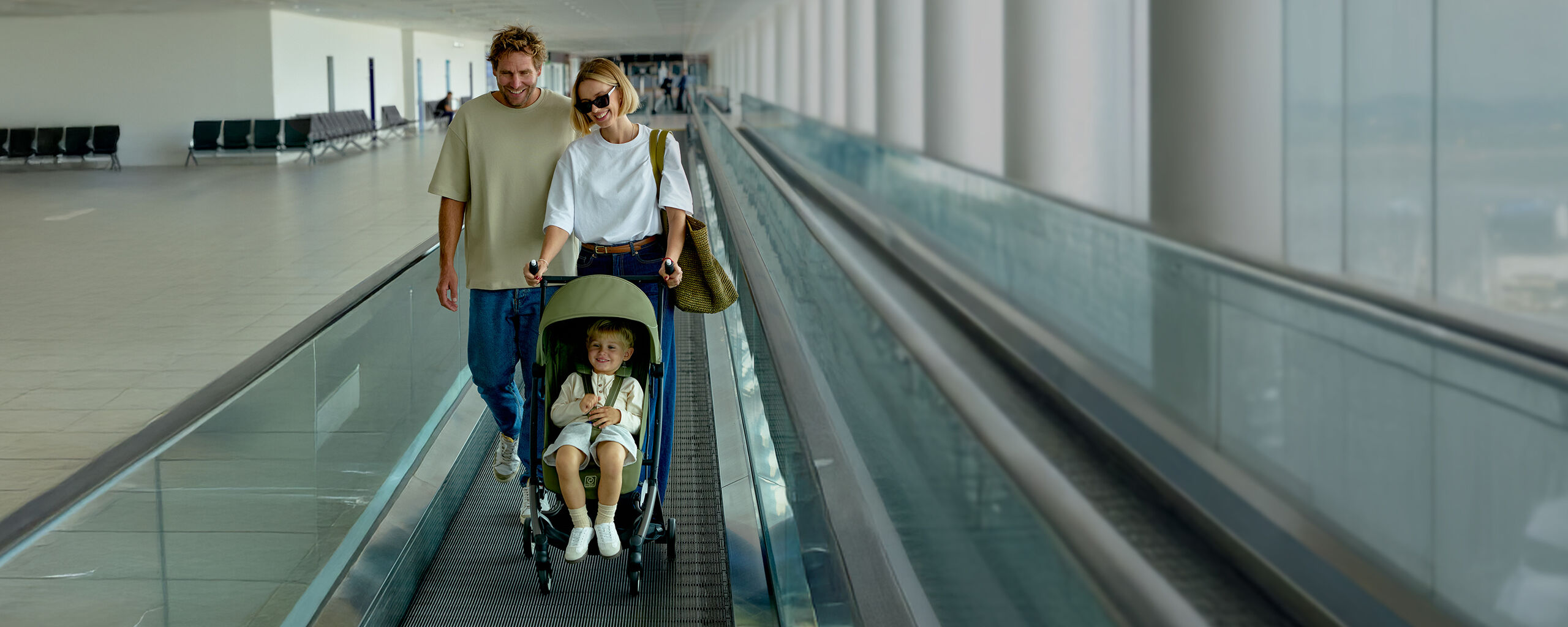 A family with a toddler in a stroller is walking on a moving walkway in a modern airport.