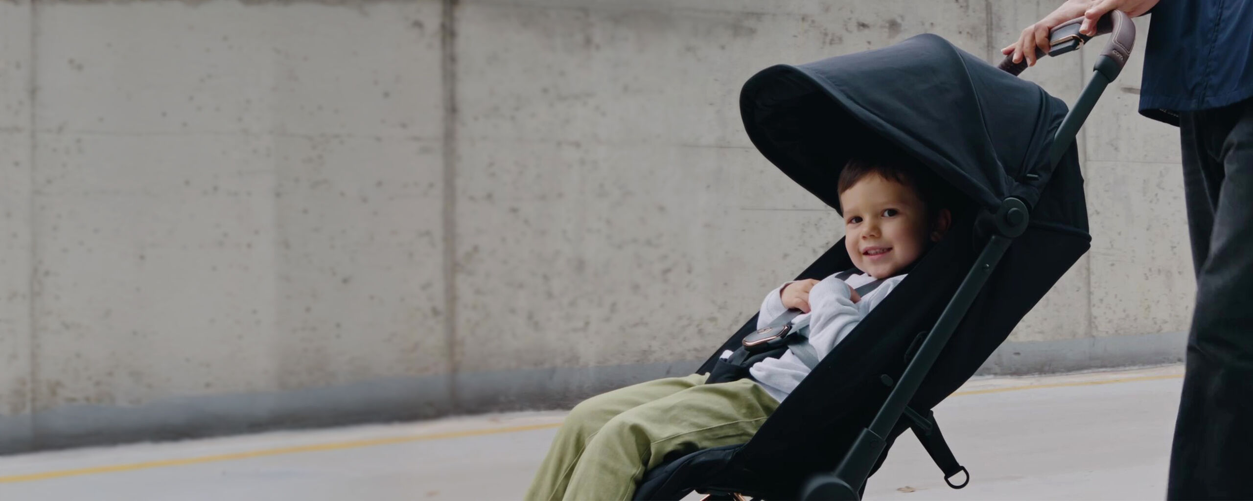 A smiling child is sitting in a black stroller with a large sunshade, while a person pushes the stroller.