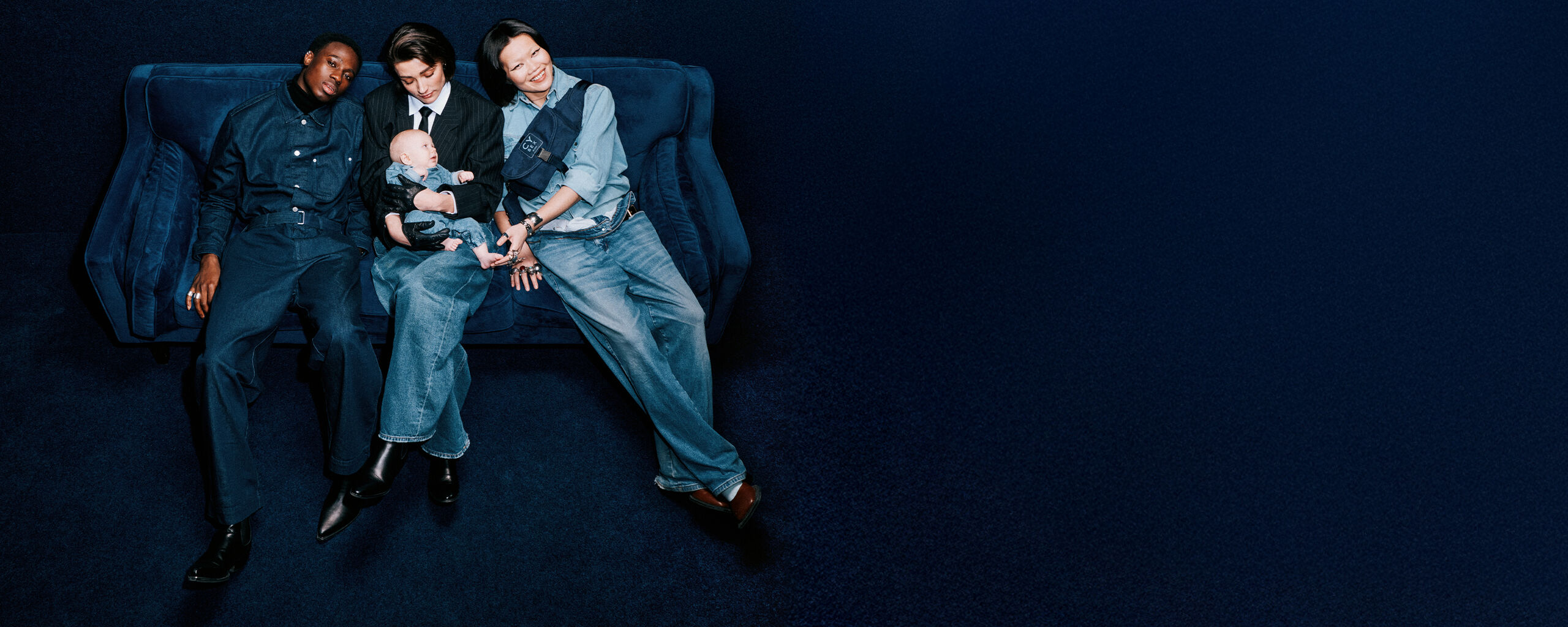 A man and two women sitting on a denim couch, the woman in the middle holding a baby in her lap, the woman on the right wearing a Rebellious Luxury Belt Bag around her.