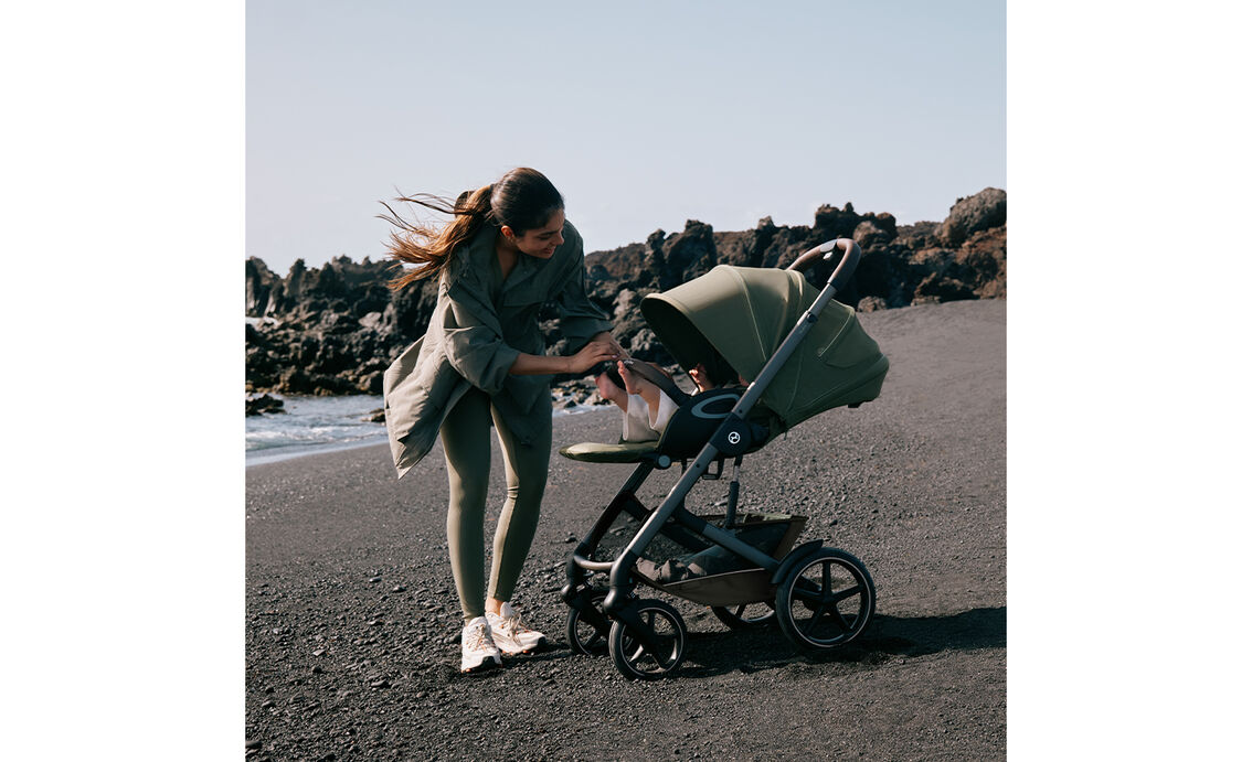 A mother checks on her baby in a Talos S Lux with the seat reclined. They're standing on a stony beach next to the ocean.