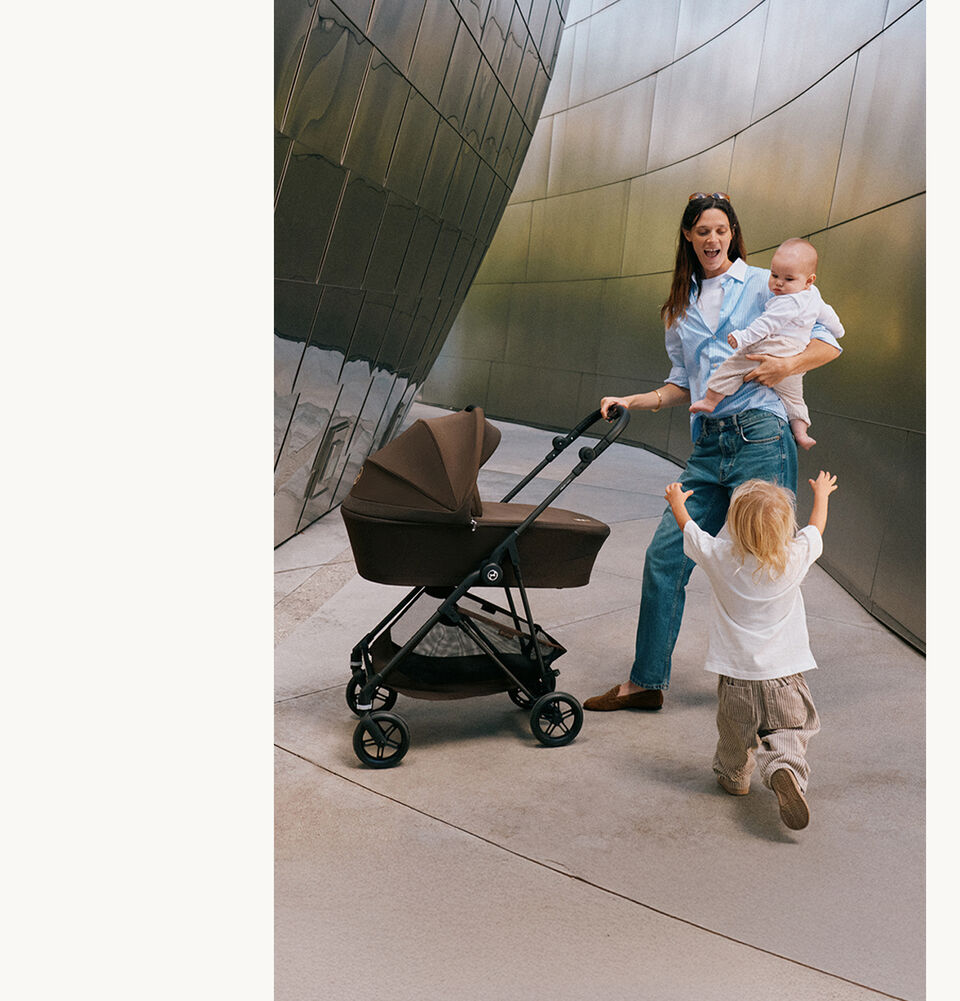 A mother with one hand on the handlebar. She's holding her toddler on her hip while her other child runs towards her with her arms up.