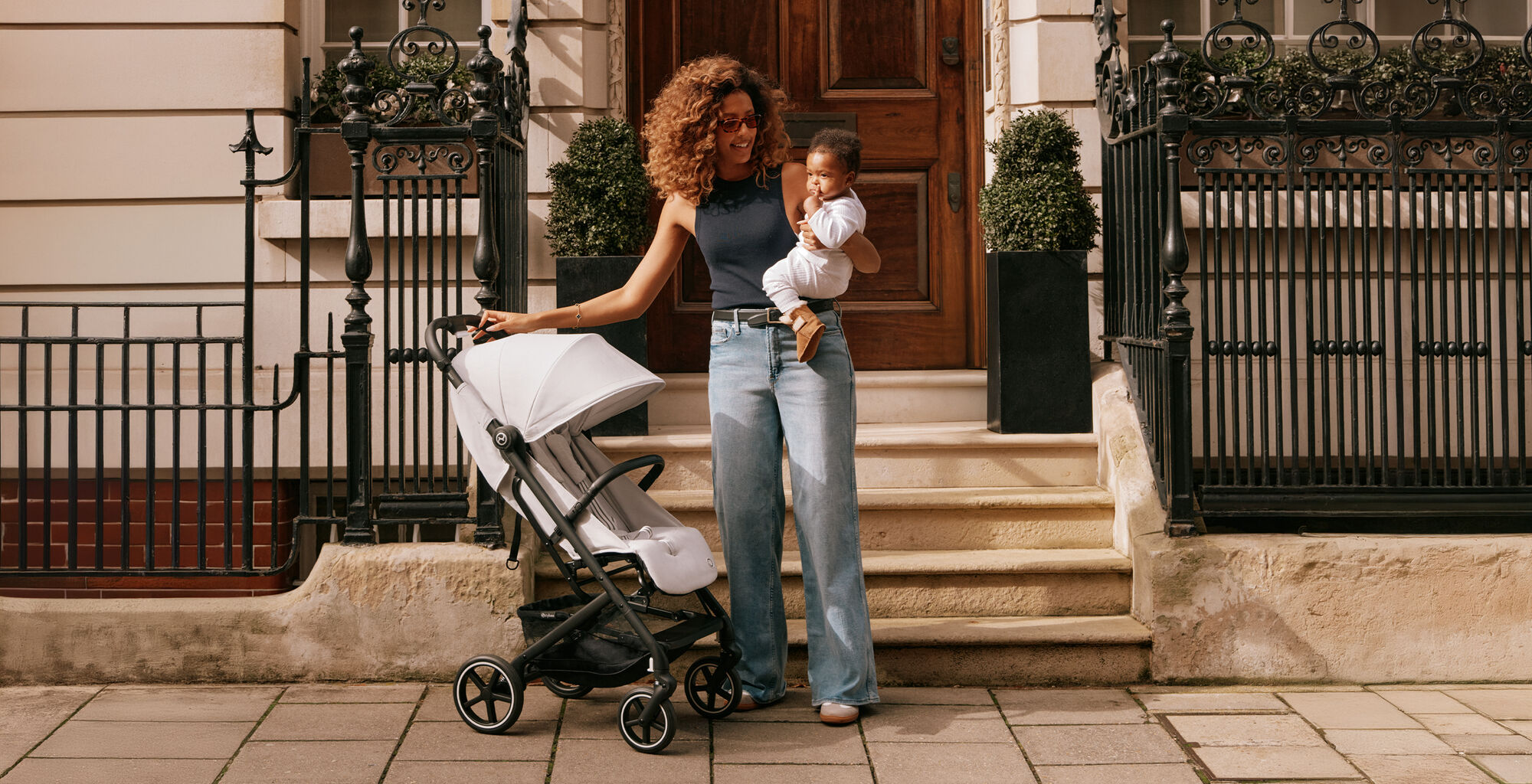 Žena stoj&iacute; s d&iacute;tětem v n&aacute;ruč&iacute; vedle koč&aacute;rku Beezy s velk&yacute;mi koly před elegantn&iacute;mi dveřmi.