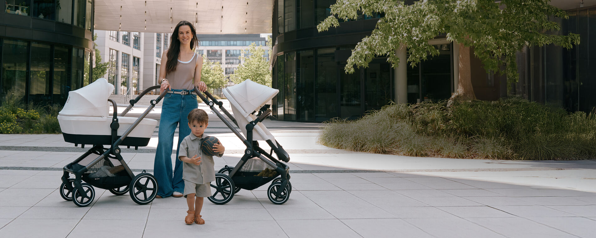Brunette mother with two CYBEX EOS Lux strollers outdoors in a summery urban scene – one stroller with seat unit, one with carrycot in off-white fabric, toddler boy with ball in foreground