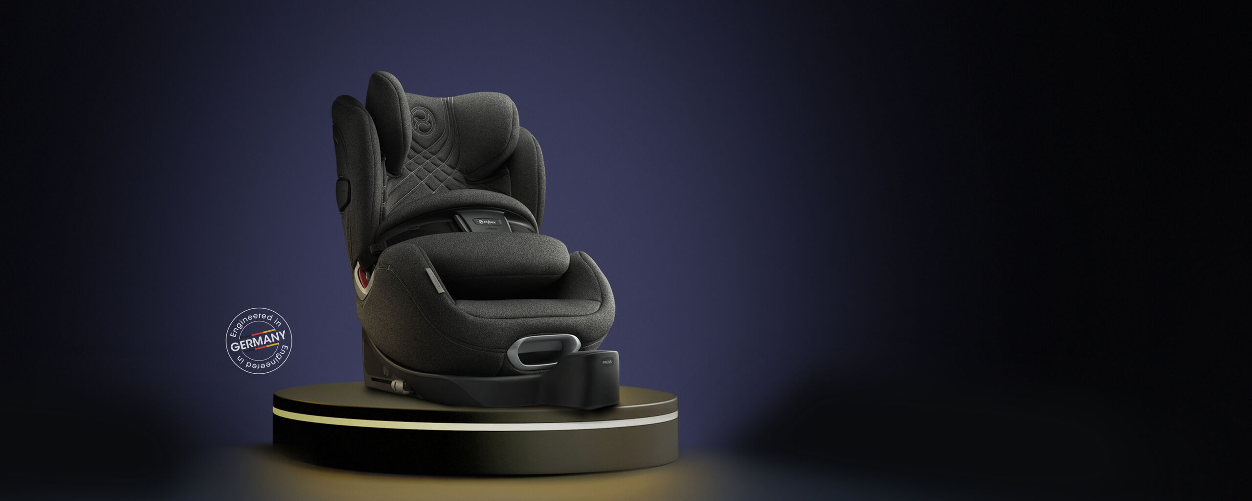Anoris T car seat in dark grey on a pedestal, with an "Engineered in Germany" emblem in the background.