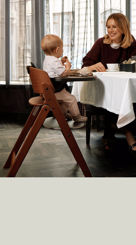 A mother sitting at a table next to her child in a Click & Fold high chair, showing him something on a piece of paper.