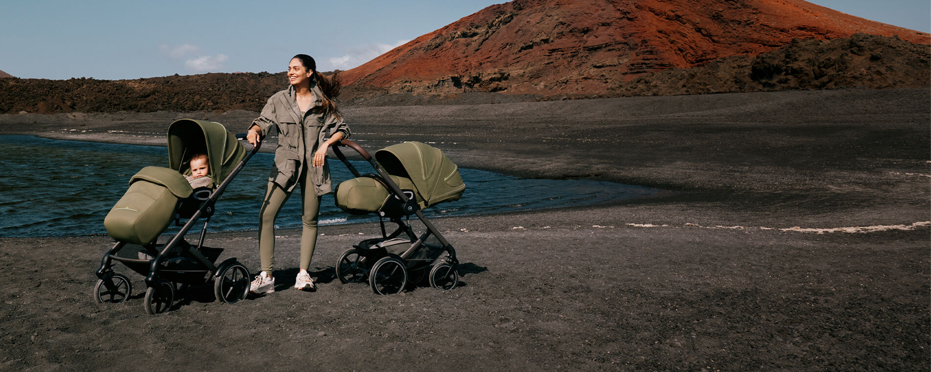 A woman standing on a stony beach with two Talos S Lux strollers, the ocean behind her.