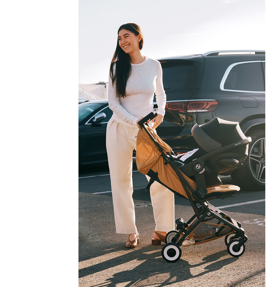 Een vrouw op een parkeerplaats buiten, voor twee auto&rsquo;s. Ze houdt een Libelle-buggy vast waaraan een autostoel voor baby's is bevestigd in het verstelbare stoel-pakket.