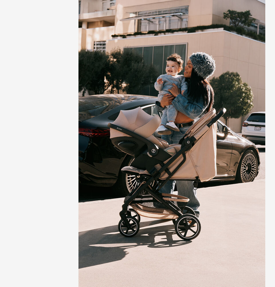 A woman holding her baby with a Beezy buggy and an infant car seat attached to it.