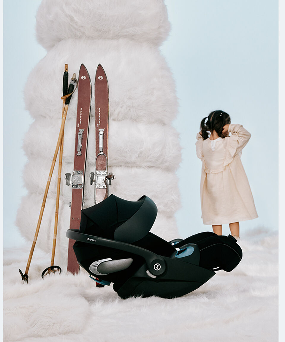Three children playing on a white rug that looks like snow. On the left, there is a CYBEX infant car seat next to them.