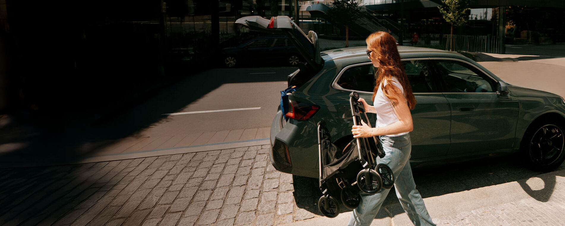 Brünette Frau mit langen Haaren steht neben einem Auto mit geöffnetem Kofferraum und hält einen zusammengeklappten Kinderwagen in der Hand.