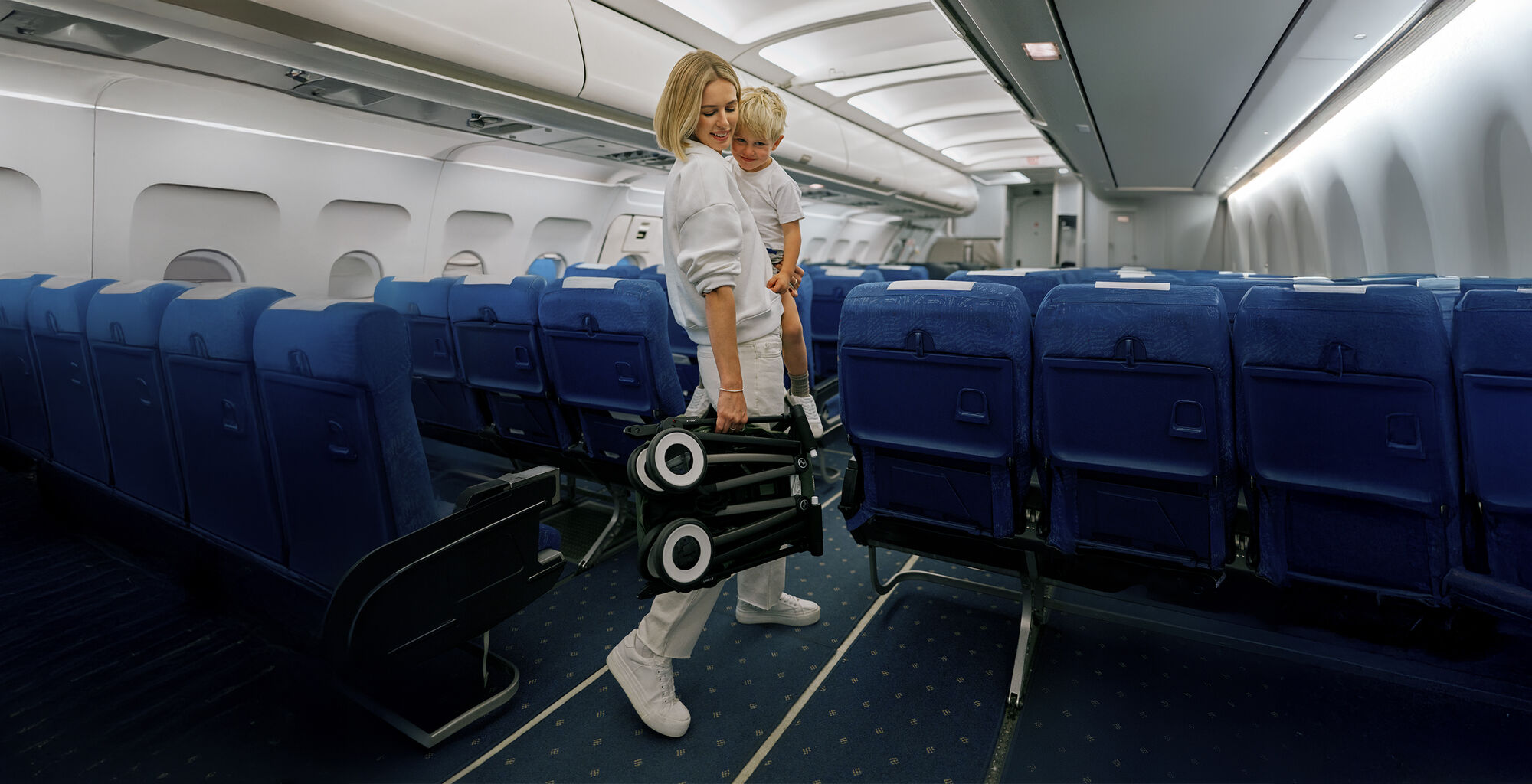 A mother is carrying a child while holding a collapsible stroller in an airplane with blue seats.