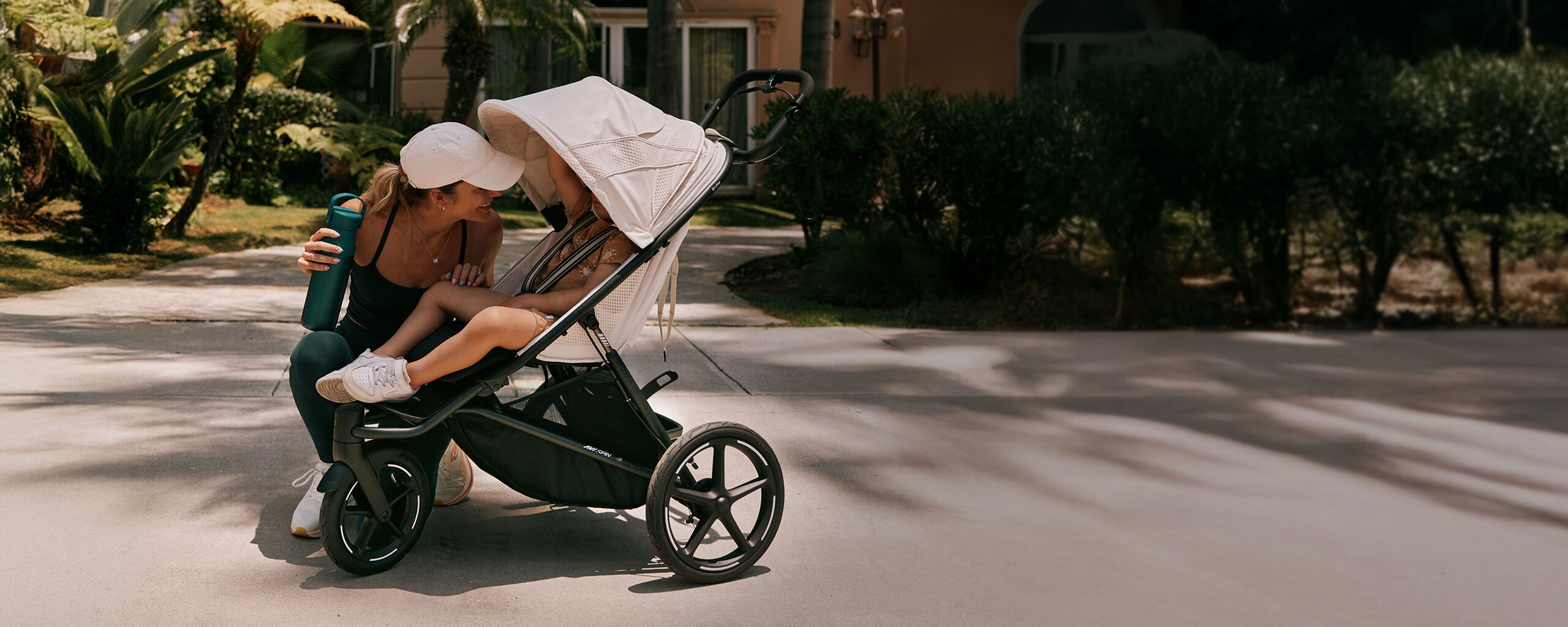 Una madre si inginocchia accanto a suo figlio in un passeggino, sorridendo mentre tiene una bottiglia d'acqua, circondata da piante verdi