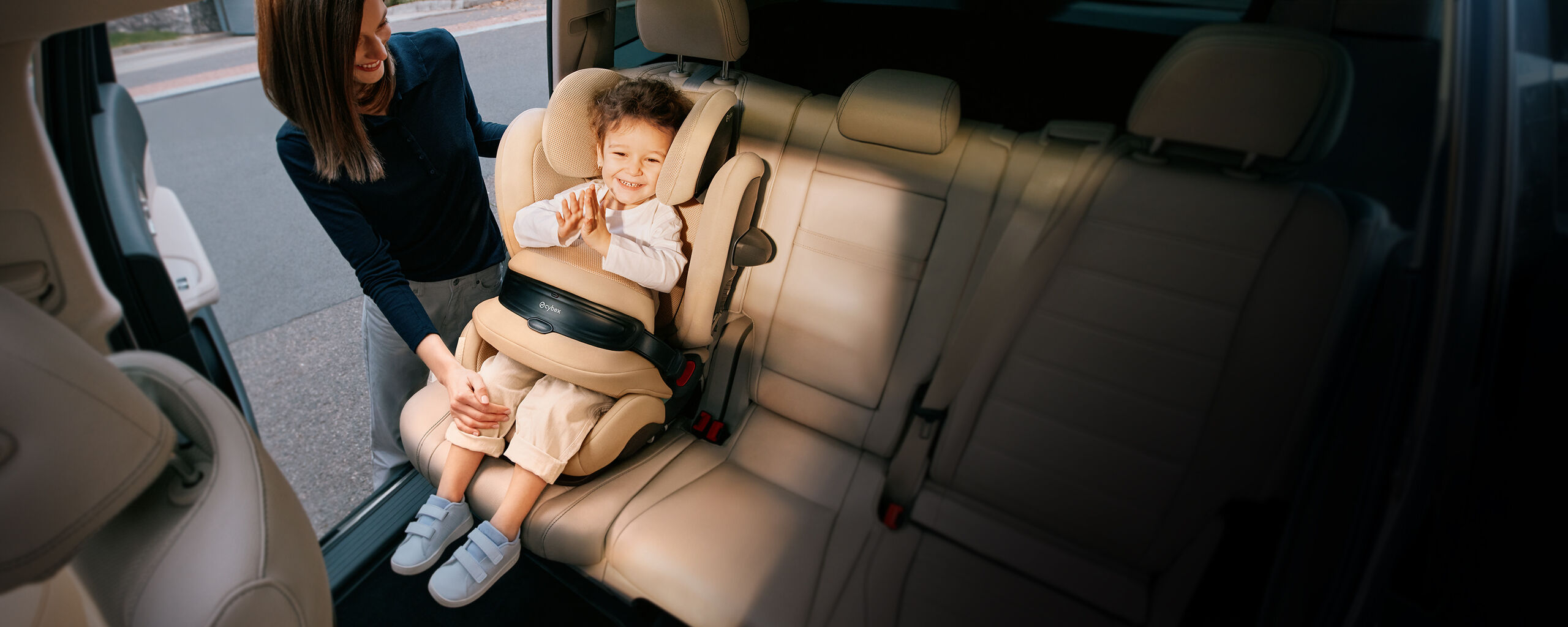 A smiling child is sitting in a Pallas G2 car seat, while a woman assists him with the seatbelt.