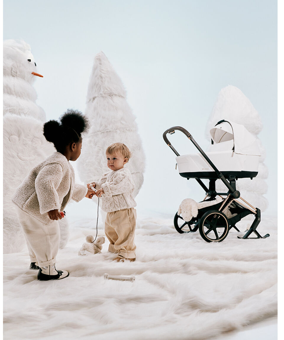 A little girl and boy playing with a toy. Behind them are makeshift white Christmas trees and a Priam with skis attached.
