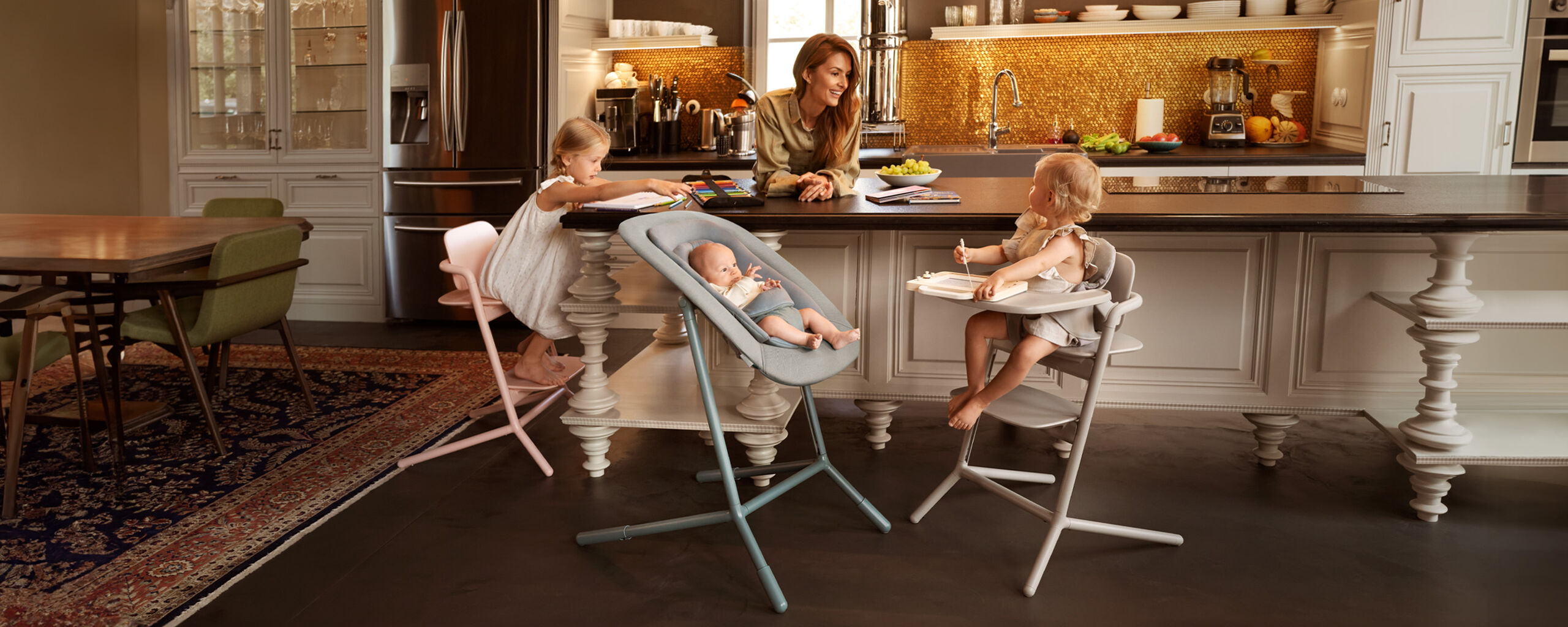 A mother sits with her three young children in a stylish kitchen. Two of the children are seated on high chairs, while the baby lies in a Lemo Training Tower.