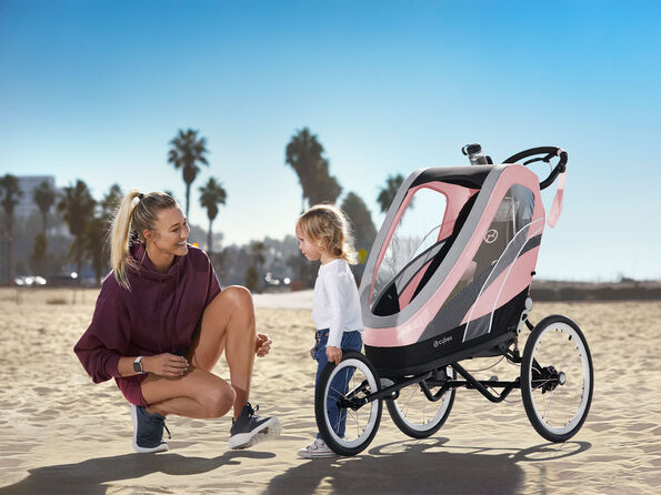 A woman kneels on the beach beside a young child and a pink CYBEX Zeno sports pushchair under sunny skies