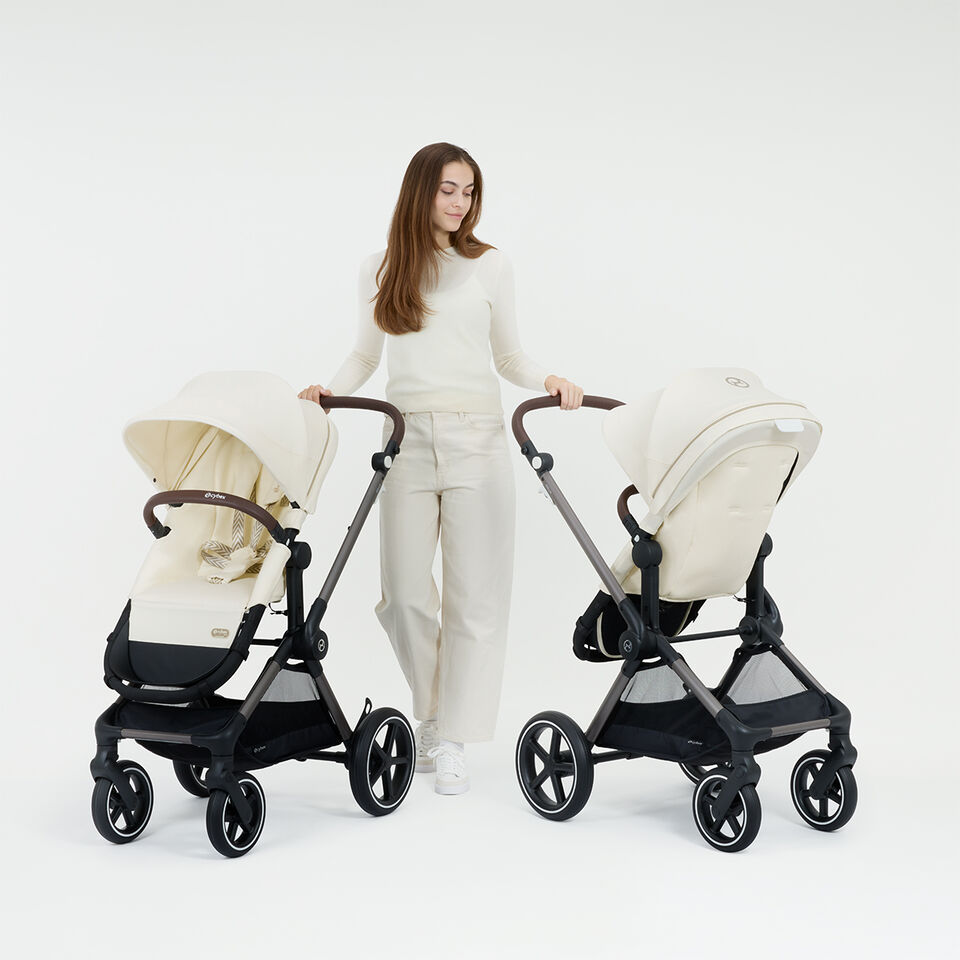 A woman standing next to two Eos Lux strollers, one with a forward facing seat unit, one with a rear facing one.