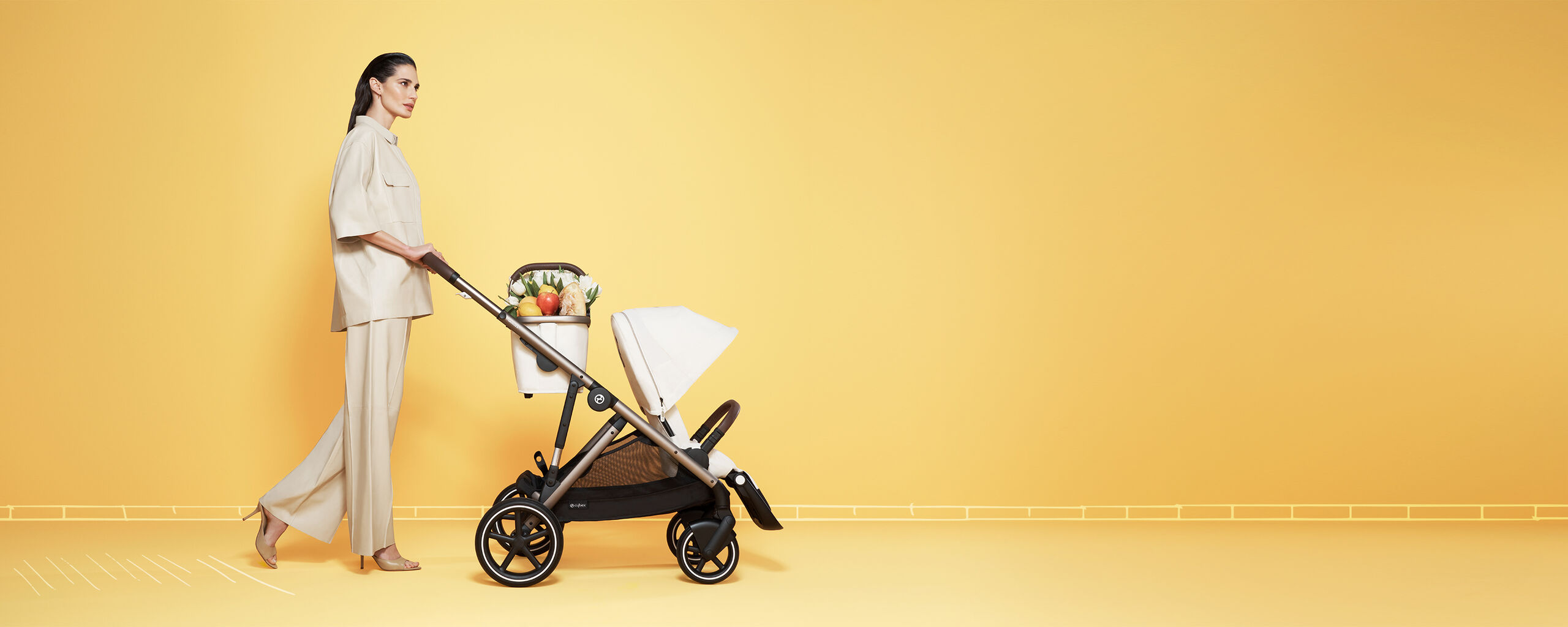 Woman in beige clothing pushing a pram with fruits and flowers against a yellow background.