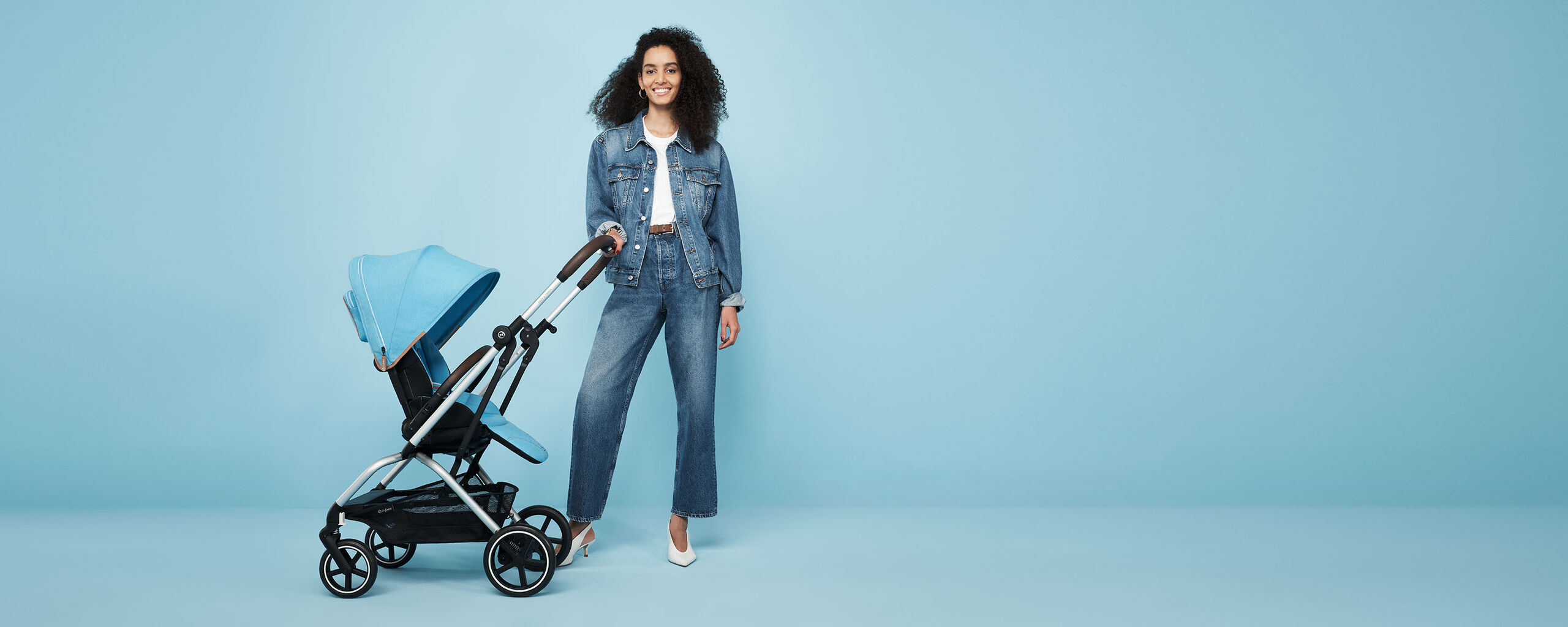 A woman in denim clothing stands next to a blue Eezy S Twist +2 pushchair against a light blue background.