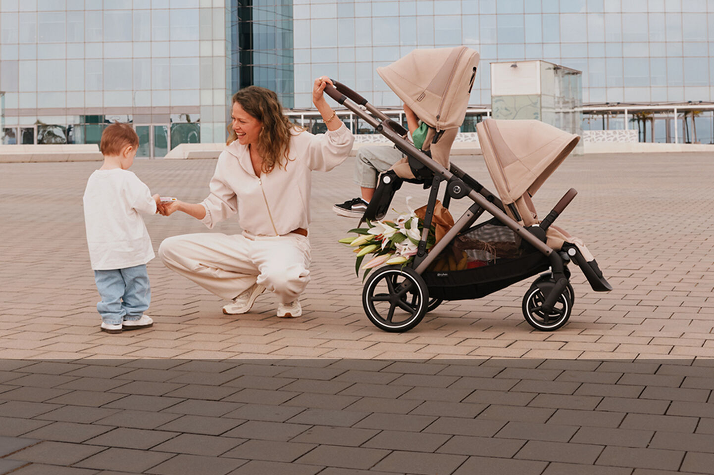 A woman laughing as she kneels down and hands something to her son, one hand on the Gazelle S stroller's handlebar with one child sitting in one of the seat units attached.