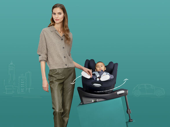 A woman stands next to a baby in a car seat against a turquoise background.