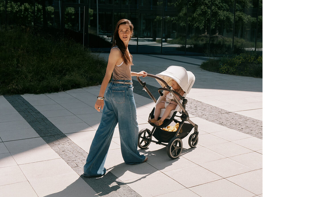 A brunette woman pushes the CYBEX EOS LUX stroller in Seashell Beige with a toddler seated inside, captured in an outdoor campaign shoot.