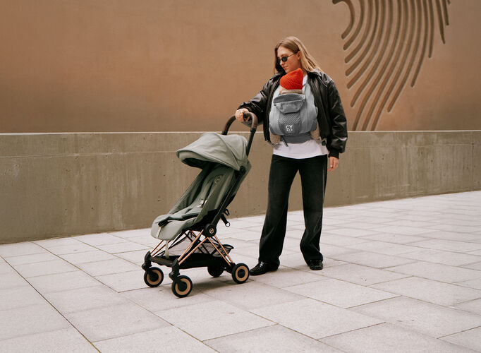 A woman with a baby carrier wearing her baby against her chest, one hand on a Coya lightweight stroller.