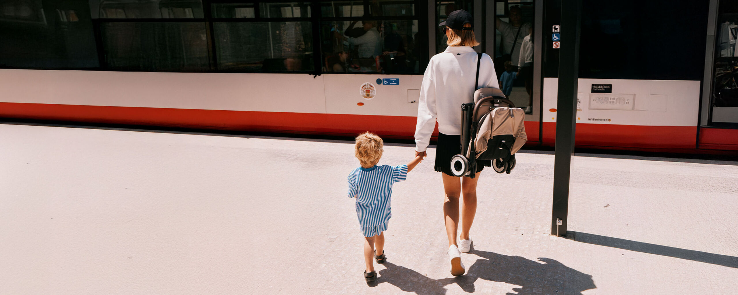 Junge blonde Frau mit Bobfrisur tr&auml;gt einen zusammengeklappten CYBEX Kinderwagen &uuml;ber der Schulter, w&auml;hrend sie die Hand eines kleinen Jungen in der N&auml;he eines Busses h&auml;lt.