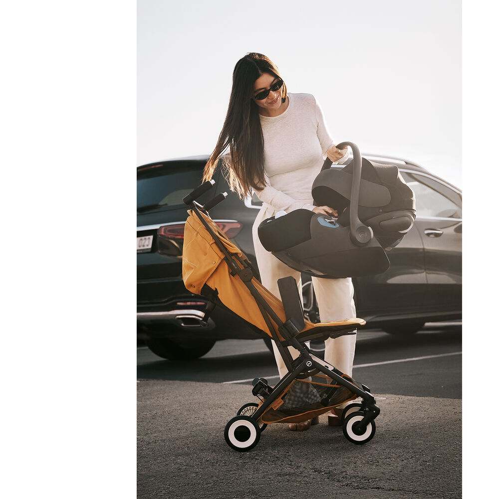 A woman in an outside parking lot in front of two cars. She's holding on to a Libelle buggy with an infant car seat attached in the recline seat pack.