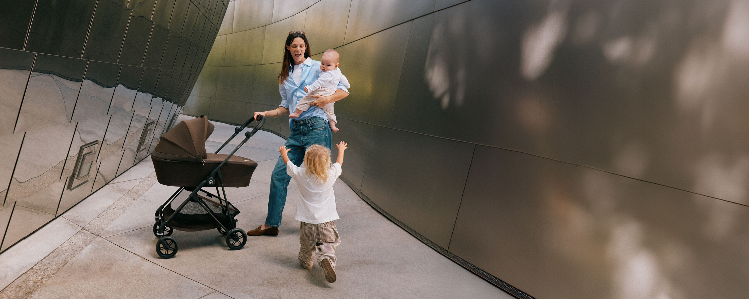 A mother holding the handlebar of her CYBEX stroller with one hand while carrying her toddler in her other arm. Her son is running towards her with open arms.