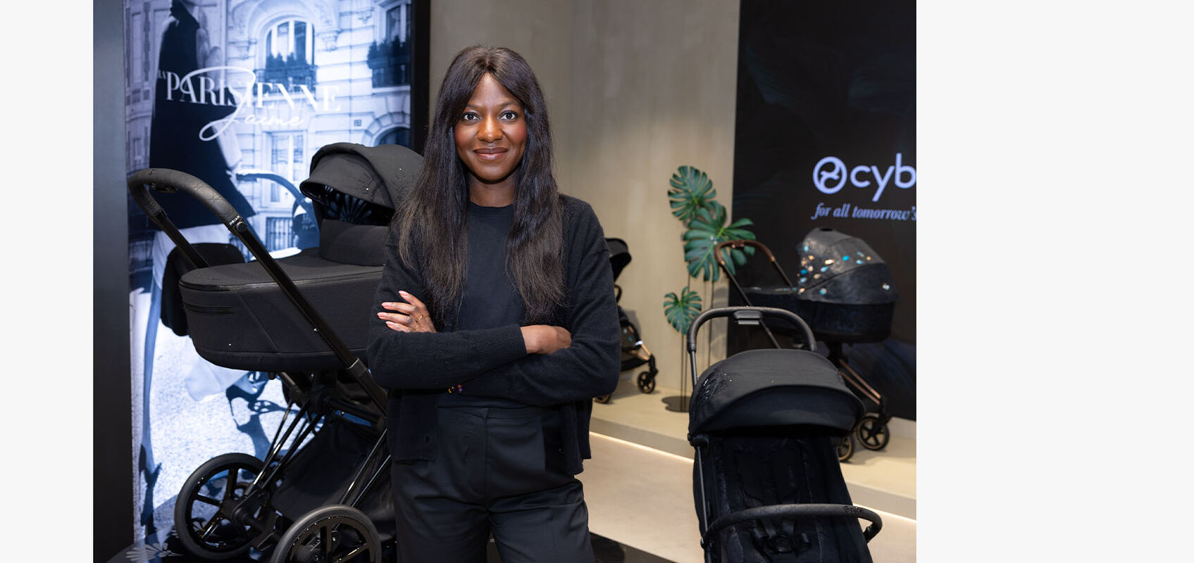CYBEX store interior: friendly-looking woman with long black hair and dressed in all black stands with arms crossed inside the store. In the background, a La Parisienne pushchair is visible.