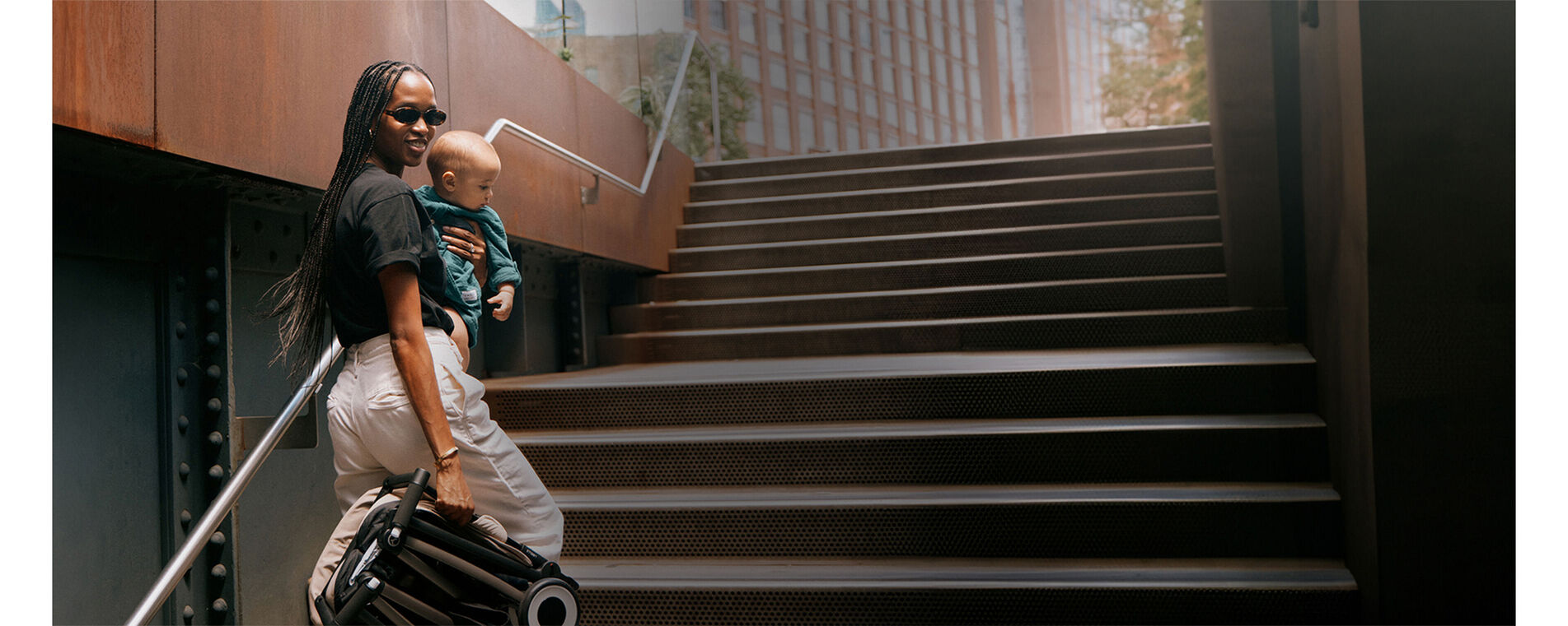 A woman walking up some stairs, her baby in her left arm, a folded up Libelle lightweight stroller in her right hand.
