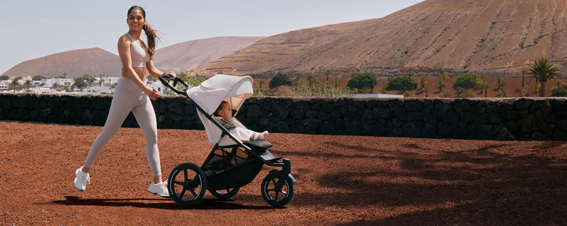 A woman in workout clothes strolling over uneven terrain with her Avi Spin and her child.