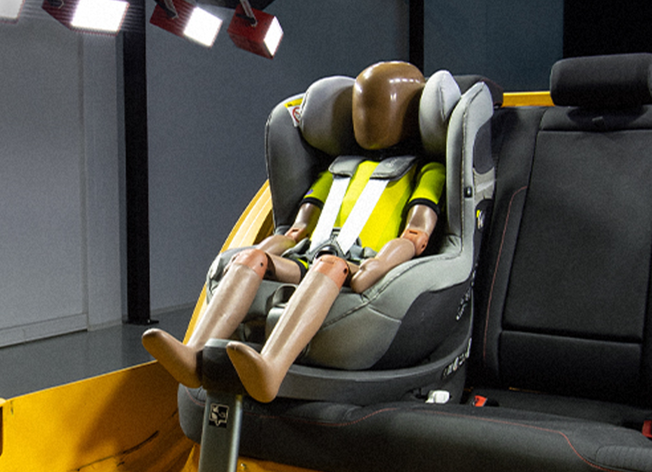 A crash test dummy is seated in a child car seat on the backseat of a yellow car frame.