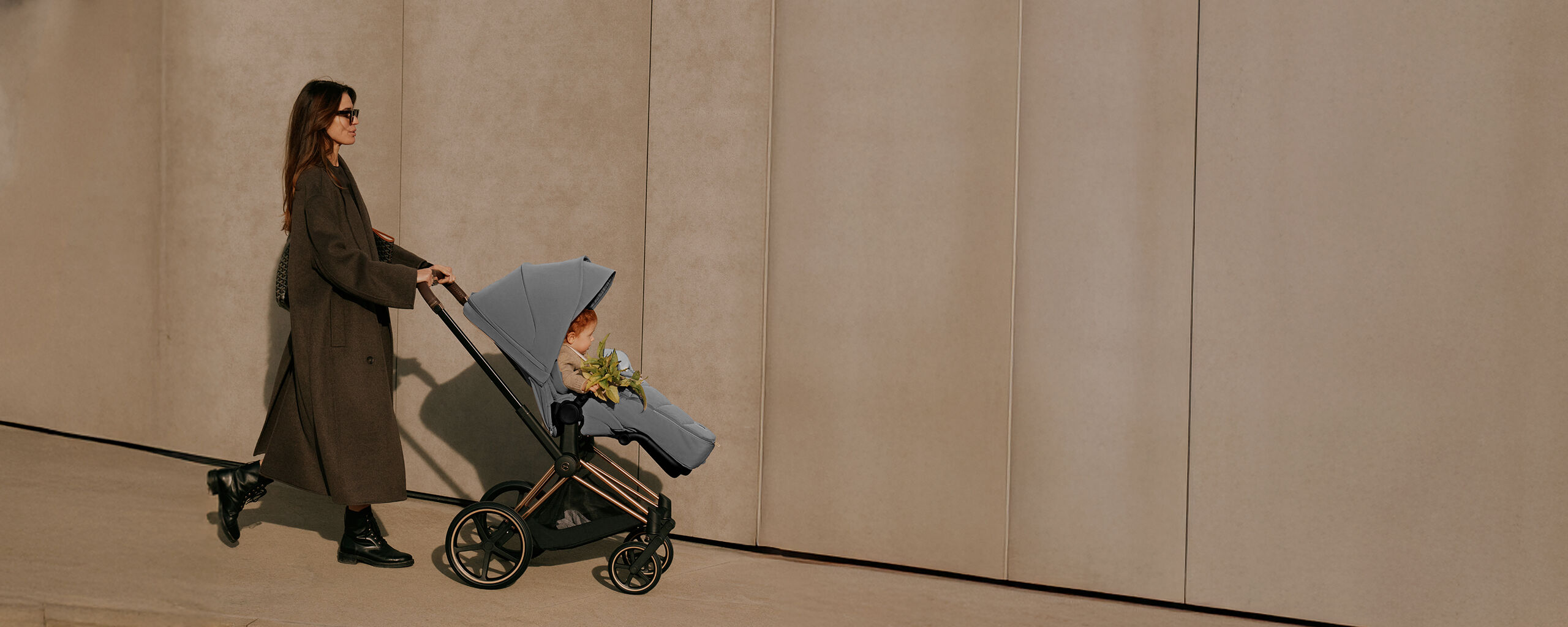 A mother wearing sunglasses pushing a CYBEX stroller with her child inside a footmuff.