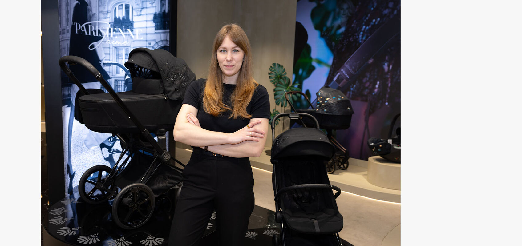 CYBEX store interior: woman with reddish-brown, loose hair dressed in black stands with arms crossed inside the store. In the background, a La Parisienne pushchair is visible.